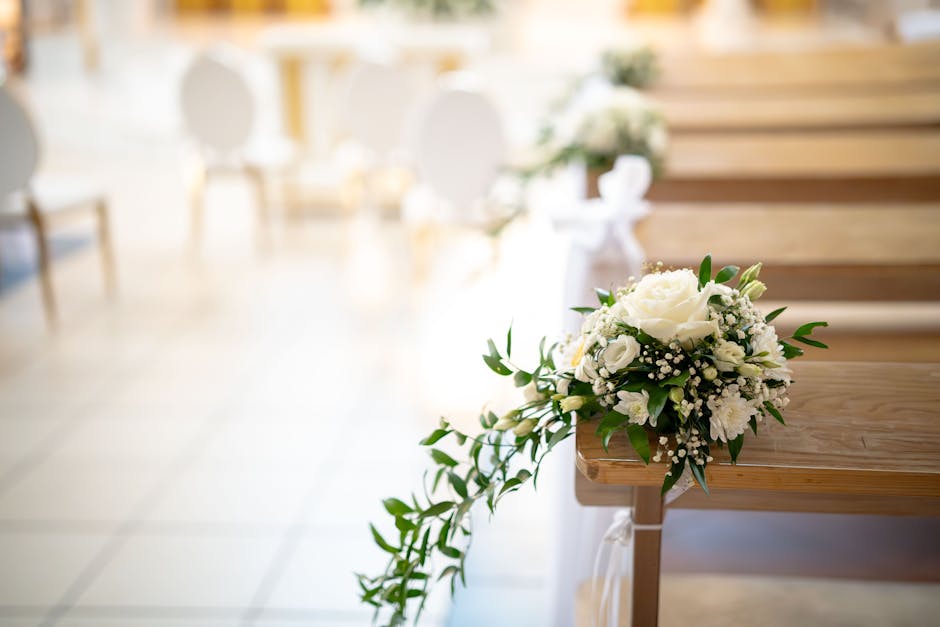 A close-up of a floral bouquet placed on a wooden pew in a softly lit indoor ceremony setting. The bouquet features creamy white roses, delicate white carnations, and sprigs of baby's breath, accented