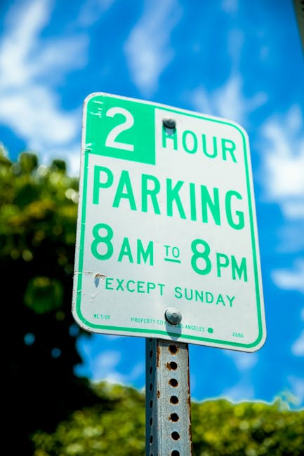 A rectangular green and white parking sign indicating a two-hour parking limit from 8 am to 8 pm, with smaller print stating 'except Sunday,' mounted on a metal post with visible rust spots. The backg