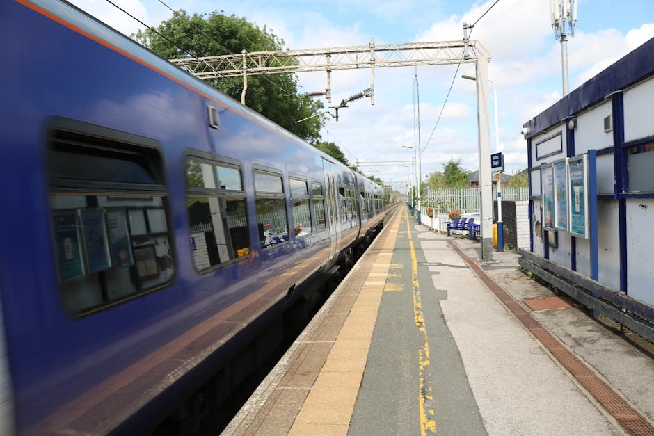 A blue train is stationary at a suburban railway platform, with its reflective windows showing the surrounding environment. The platform features a beige and gray tiled surface, with yellow safety lin