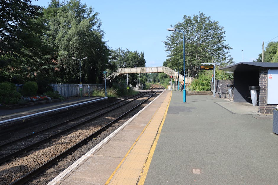 A quiet train station platform at South Hornchurch with two parallel railway tracks running alongside a paved waiting area. The platform is bordered by a yellow safety line and features a blue lamppos