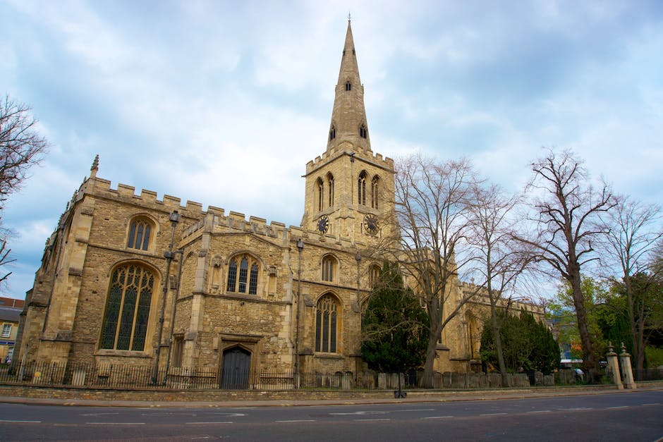 A tall, historic stone church with a pointed spire and large stained glass windows, situated along a street with leafless trees in the foreground. The church features Gothic architectural elements, in