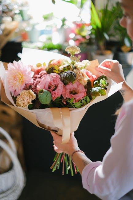 A person holding a vibrant bouquet of fresh flowers, including pink peonies, white and pink roses, and small purple and white filler flowers, wrapped in beige and white paper with a cream ribbon. The 