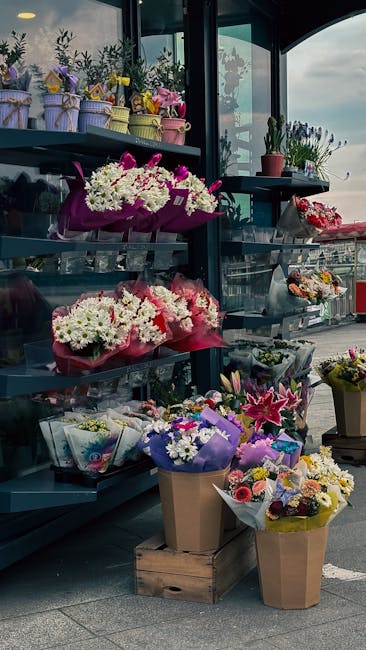 A display of various fresh flower arrangements outside a floral shop, featuring bouquets wrapped in colorful paper and placed on shelves and the ground. The arrangements include a mix of white, pink,